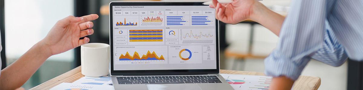 Two people point at a laptop displaying business charts and graphs on a desk with a white mug and papers.