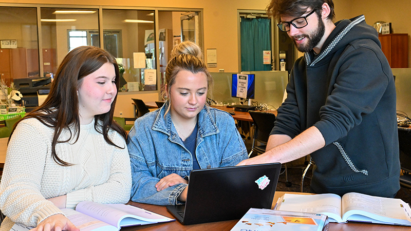 Three students studying at a table. Two women are seated, examining a laptop, while a man stands beside them, pointing at the screen.