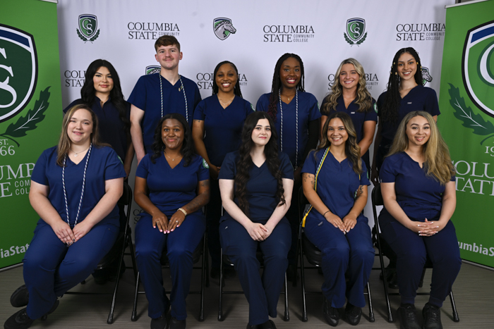 A group of people sit and stand in front of a white and green background. 
