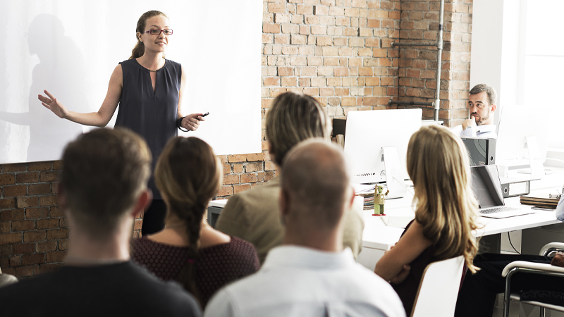 A woman in a dark sleeveless dress speaks to an audience in a brick-walled office, standing beside a projected screen with coworkers seated at desks.