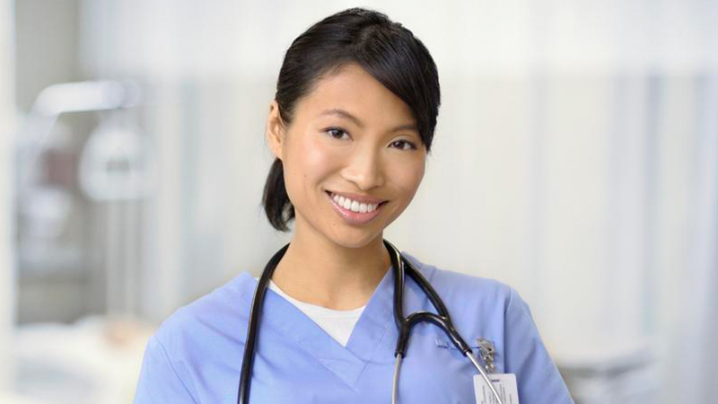 A smiling female healthcare worker in blue scrubs with a stethoscope around her neck stands in a bright medical setting.