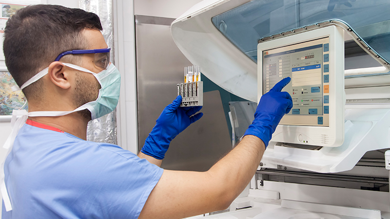 A masked lab technician in blue gloves operates a touchscreen analyzer while holding a test tube rack in a clinical laboratory.